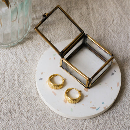 A small open jewelry box with a brass metal frame and clear glass lid sits on a round white speckled coaster. Two gold hoop earrings rest on the tray beside the box.