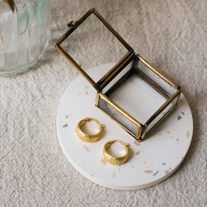 A small open jewelry box with a brass metal frame and clear glass lid sits on a round white speckled coaster. Two gold hoop earrings rest on the tray beside the box.