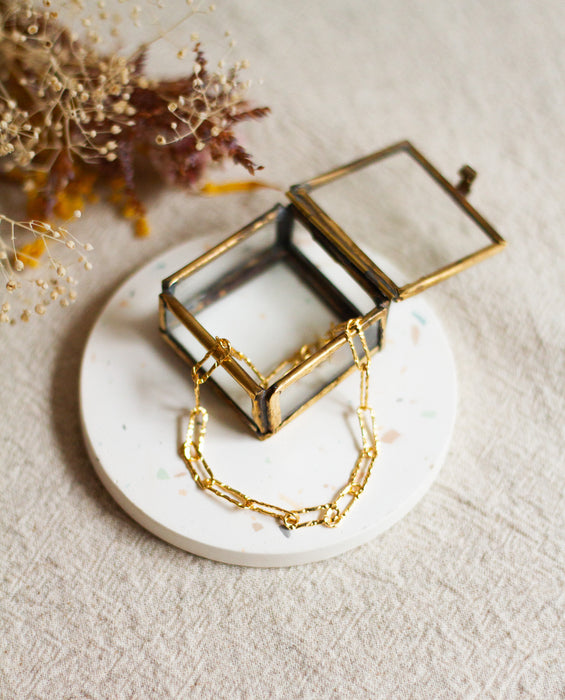 Gold hammered link bracelet on a white dish with a glass boxes and dried flowers on a beige linen surface