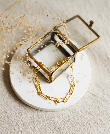 Gold hammered link bracelet on a white coaster with dried flowers and a glass box on a beige linen background