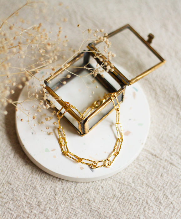 Gold hammered link bracelet on a white coaster with dried flowers and a glass box on a beige linen background