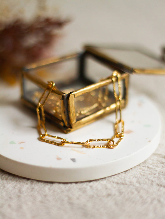 Gold hammered link bracelet in a small glass and brass box on a white coaster with a blurred background