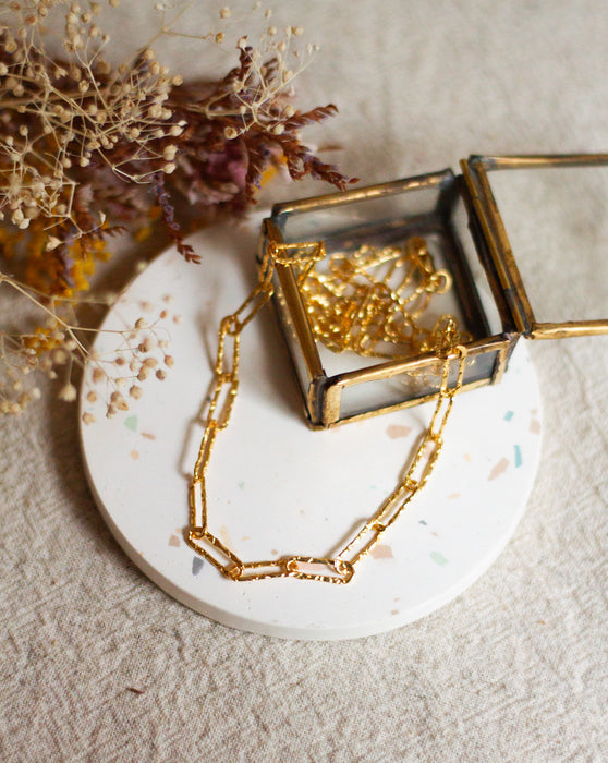 Gold hammered link necklace on a white coaster with dried flowers on a beige linen background