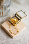Gold hammered link necklace on a wooden block in an open glass jewellery box with a beige linen background