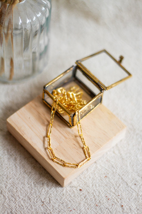 Gold hammered link necklace on a wooden block in an open glass jewellery box with a beige linen background