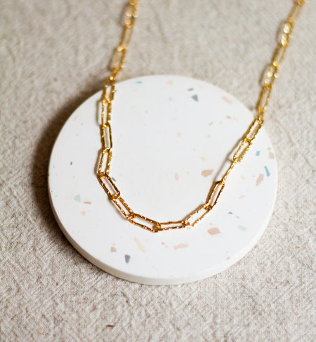 Gold hammered link necklace on a round white coaster and a beige linen surface