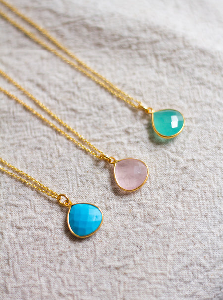 Three gold necklaces with muted teardrop shaped pendants on a neutral linen surface