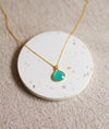 Gold necklace with an aqua pendant on a round, speckled coaster with a beige linen background
