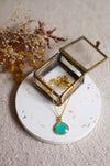 Gold necklace with an aqua pendant in an open glass  jewellery box on a linen surface with dried flowers.