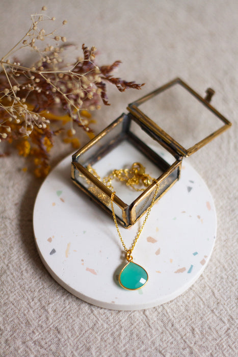 Gold necklace with an aqua pendant in an open glass  jewellery box on a linen surface with dried flowers.