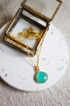 Gold necklace with an aqua teardrop pendant in an open glass jewellery box on a white coaster surface with a beige linen background.