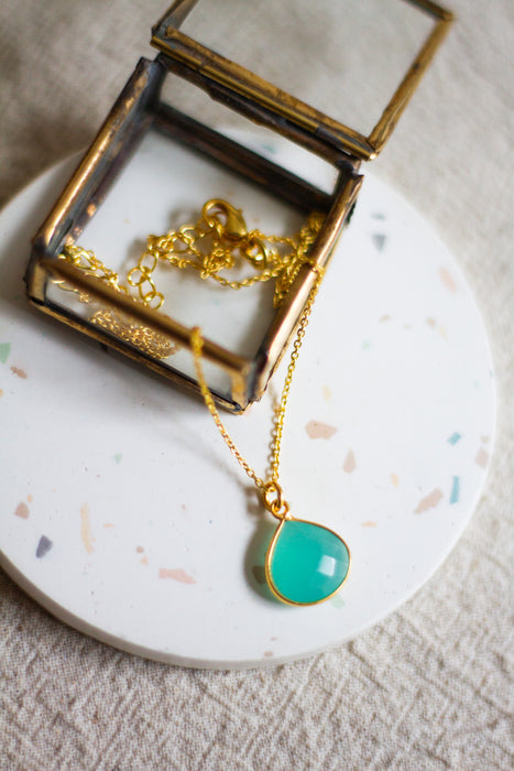 Gold necklace with an aqua teardrop pendant in an open glass jewellery box on a white coaster surface with a beige linen background.