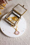 Gold necklace with a rose quartz pendant in an open glass jewellery box on a linen surface.