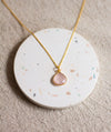 Gold necklace with a rose quartz teardrop pendant on a round, speckled coaster, and a beige linen background.