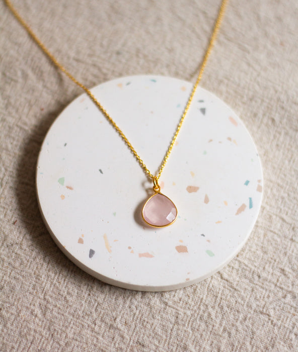 Gold necklace with a rose quartz teardrop pendant on a round, speckled coaster, and a beige linen background.