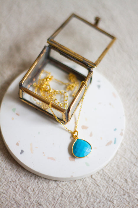 Gold necklace with a turquoise teardrop pendant in an open glass jewellery box on a linen surface