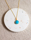 Gold necklace with a turquoise pendant on a round, speckled coaster with a beige linen background