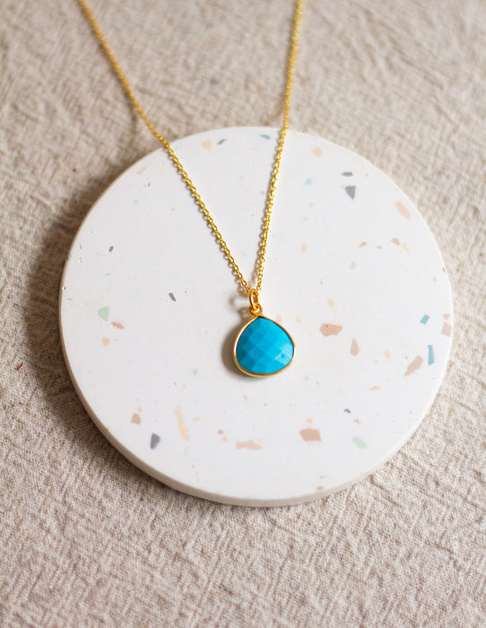 Gold necklace with a turquoise pendant on a round, speckled coaster with a beige linen background