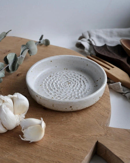 Ceramic garlic grater on a wooden surface chopping board, with garlic and wooden utensils in the background