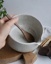 White ceramic matcha bowl being held by a hand, with wooden spoon on a wooden surface