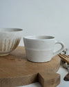 Ceramic bowl and mug with a white speckled glaze on a wooden chopping board with a neutral background