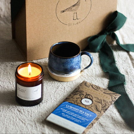 Gift set with a mug, candle, and bar of chocolate on a textured surface with a branded The Painted Bird box.