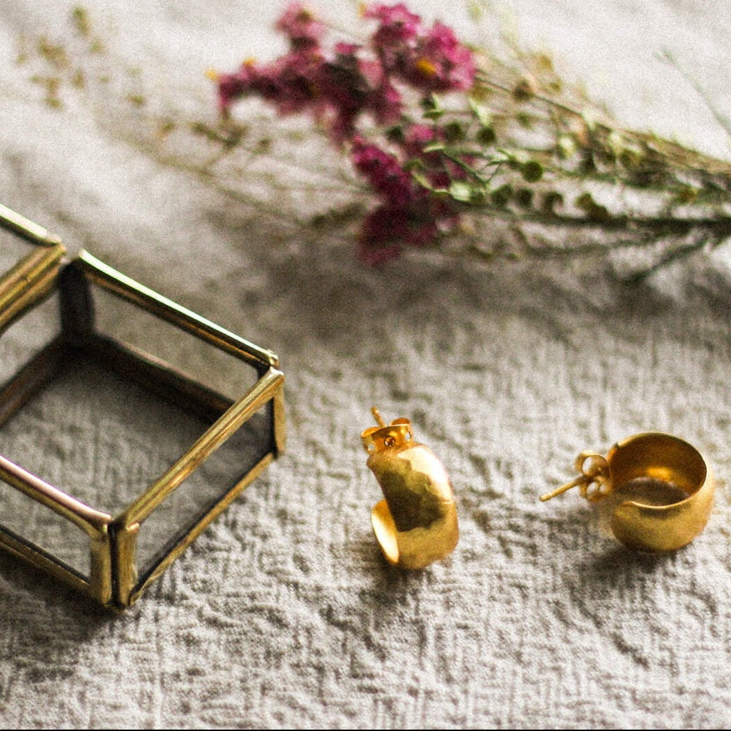 Gold earrings on a textured surface with a small glass box and dried flowers.
