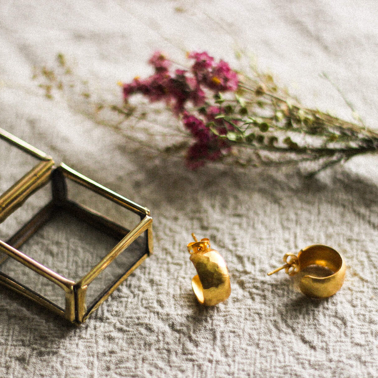 Gold earrings on a textured surface with a small glass box and dried flowers.