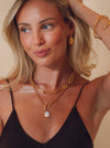 Woman wearing gold hoop earrings, and layered gold necklaces and bracelets, standing against a beige background