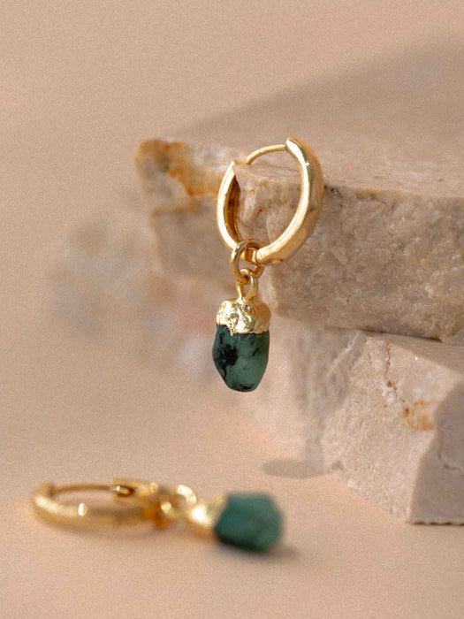 Gold hoops with emerald stones on a stone surface, with a blurred background
