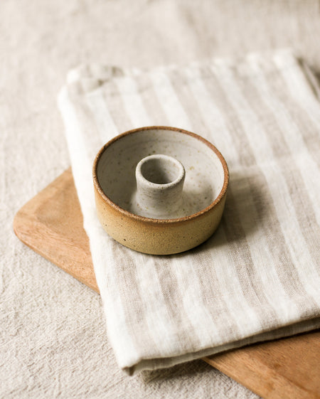 Beige ceramic candlestick holder on a wooden breadboard and beige striped tea towel, with a beige linen background 