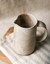 Beige ceramic pitcher on a wooden chopping board and striped tea towel with a blurred neutral background