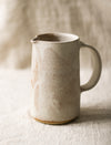 Speckled beige ceramic pitcher on a textured surface with a neutral background