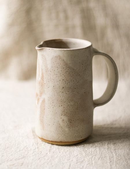 Speckled beige ceramic pitcher on a textured surface with a neutral background