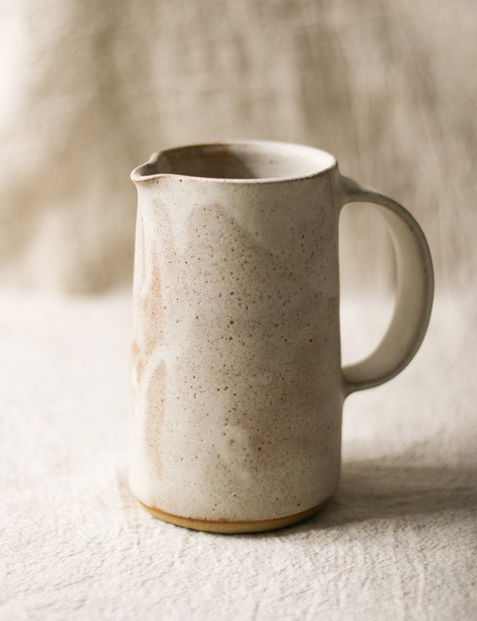 Speckled beige ceramic pitcher on a textured surface with a neutral background