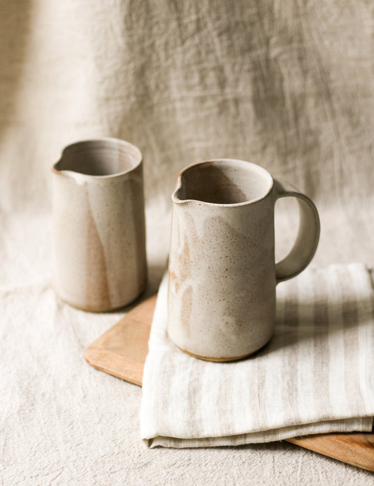Two beige ceramic pitchers on a wooden chopping board and striped tea towel with a neutral background