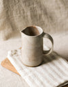 Beige ceramic pitcher on a wooden board and a beige striped tea towel with a neutral background