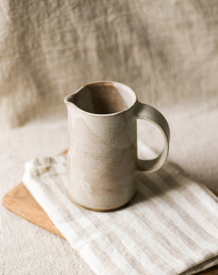 Beige ceramic pitcher on a wooden board and a beige striped tea towel with a neutral background
