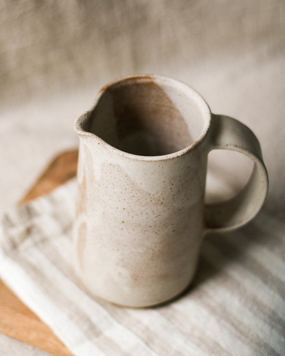 Beige ceramic pitcher on a wooden chopping board and striped tea towel with a blurred neutral background