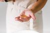 a persons hand holding a body oil in an amber galss bottle, and pouring it into the hand which is held out, with a blurred background