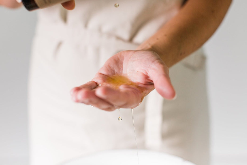 a persons hand holding a body oil in an amber galss bottle, and pouring it into the hand which is held out, with a blurred background