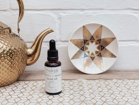 A bottle of 'Moon Facial Oil' by Moksa, placed next to a brass teapot and a geometric patterned plate on a textured surface.