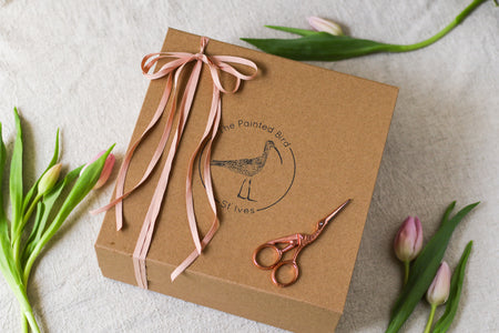 Brown kraft gift box with pink ribbon, sewing scissors, and pink tulips on a beige linen background
