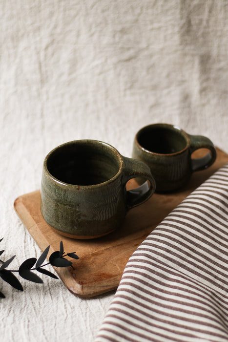 Two dark green ceramic mugs on a wooden board with a striped cloth and plant in the background.