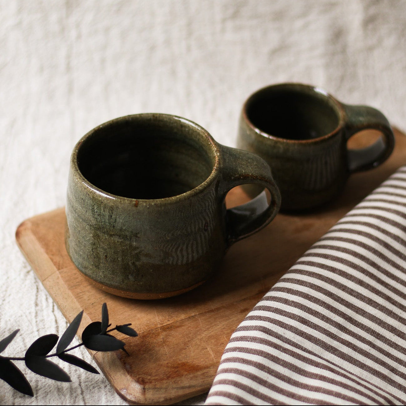 Two dark green ceramic mugs on a wooden board with a striped cloth and leaves