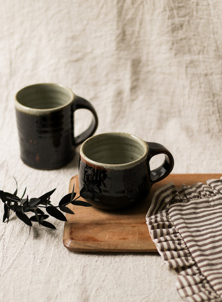 Two black ceramic mugs on a wooden board with a striped cloth and plant leaves on a textured beige background.
