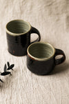 Two dark brown ceramic mugs on a beige surface with leaves in the background