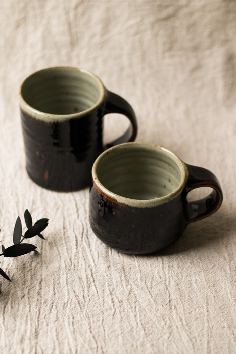 Two dark brown ceramic mugs on a beige surface with leaves in the background