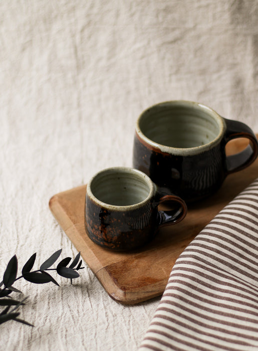Two brown ceramic mugs on a wooden board with a striped fabric and leaves in the background