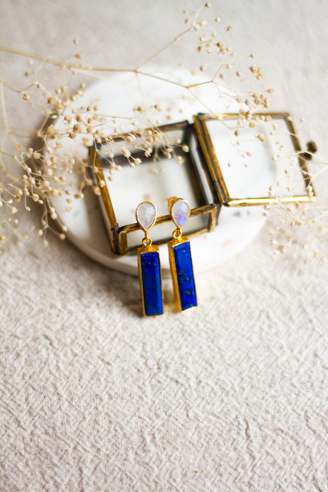 Lapis and moonstone earrings in a glass box, set on a white coaster and a linen surface